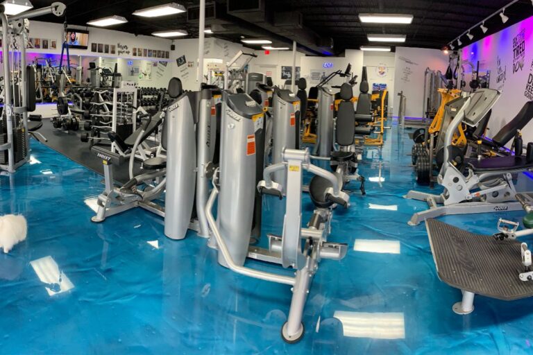 Wide-angle view of a well-equipped gym with various exercise machines, a shiny blue floor, and motivational phrases on the walls.