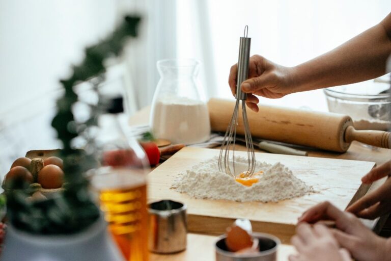 A person whisking an egg into a mound of flour on a wooden countertop with baking tools, ingredients, and rolled dough in the background.