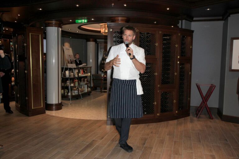 Chef Curtis Stone addressing an audience with a microphone, wearing a white chef's jacket and striped apron in an upscale restaurant setting.