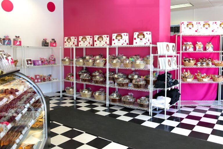 Interior of a vibrant bakery with pink walls and a checkered floor showcasing shelves full of gift baskets and treats, and a display case with pastries.