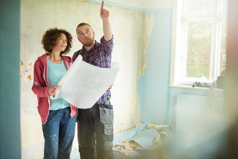 A man and woman discussing renovation plans in a room with peeling blue paint, while holding and looking at a blueprint.