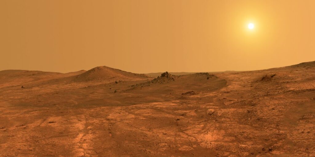 A barren, undulating Martian landscape extends under a hazy, golden sky, with the sun casting a surreal glow over the horizon.