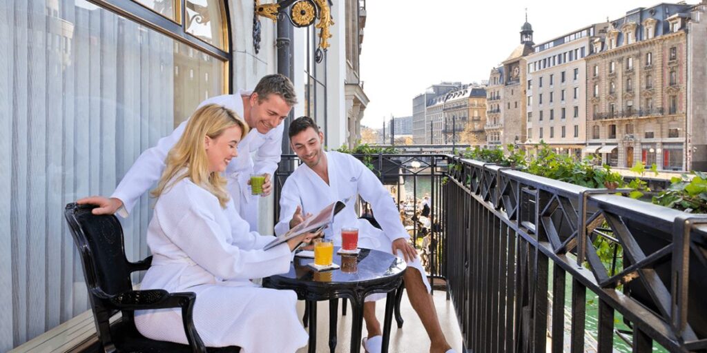 Three people in white robes enjoying beverages and reading a menu on a balcony overlooking a city street.