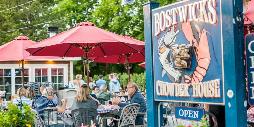 Outdoor dining scene at Bostwick's Chowder House with patrons under red umbrellas.