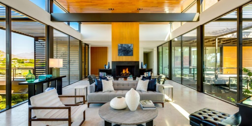 Modern living room with high ceilings, a central fireplace, and a wall of windows showcasing desert views.