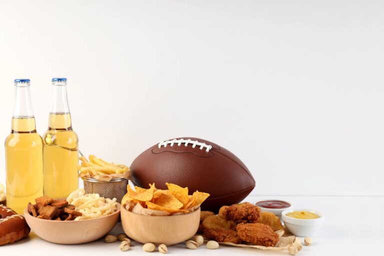 An assortment of game day snacks with a football, two bottles of soda, and a white backdrop.