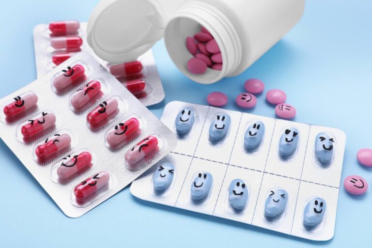 Various capsules and pills with happy and sad face emoticons printed on them, spilling from an open bottle onto a blue surface.