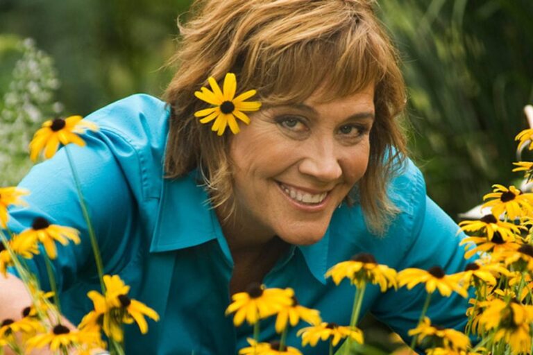 A cheerful woman with brown hair adorned with a yellow daisy flower smiling among yellow daisy flowers in a garden.