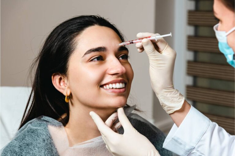 A smiling woman receiving a cosmetic injection on her forehead from a healthcare professional.