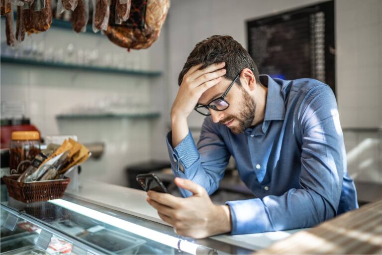 A man in a blue shirt looking stressed while checking his phone in a deli shop.
