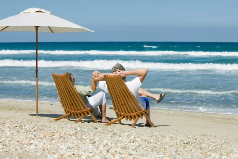 A couple relaxing on wooden lounge chairs under a white umbrella on a pebbly beach with waves in the background.