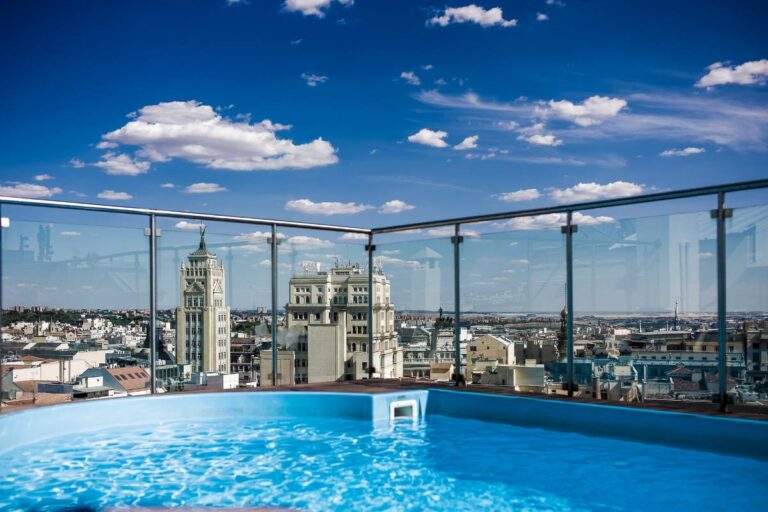 A rooftop swimming pool with a transparent glass fence offers a panoramic view of an urban skyline under a clear blue sky.