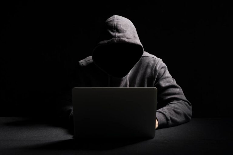 A person in a hoodie with their face obscured sitting in front of a laptop, suggesting online anonymity or cybersecurity concept.