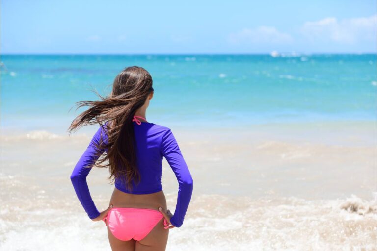 A woman standing on the beach, wearing a long-sleeved sun protective rash guard and pink bikini bottoms.
