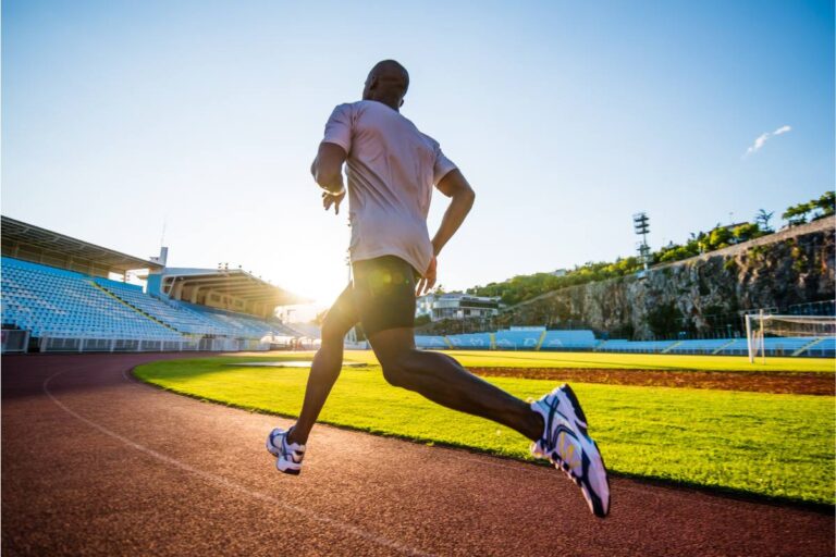 Male athlete in mid-stride running on a track in a stadium with the sun low on the horizon casting a warm glow over the scene.