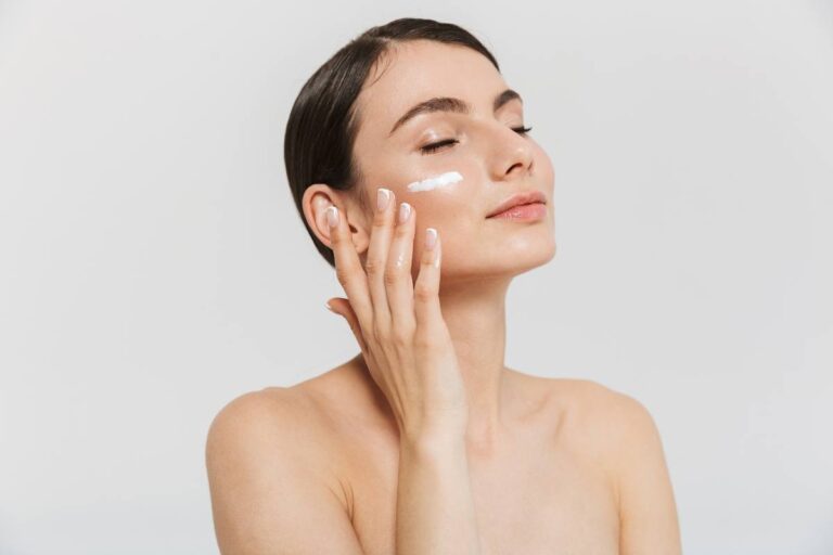 A woman with a serene expression gently applies cream to her cheek against a white background.