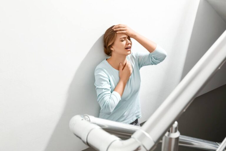 A woman in discomfort holding her throat and head while standing on stairs.
