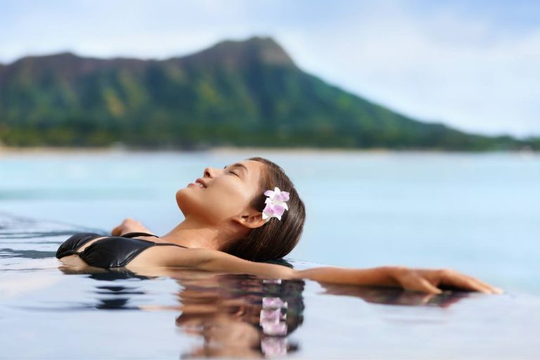 A serene woman floating in an infinity pool with a mountainous backdrop, adorned with a white flower in her hair.