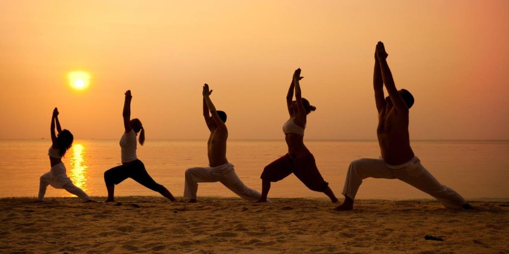 Silhouettes of five people practicing yoga on a sandy beach at sunrise.