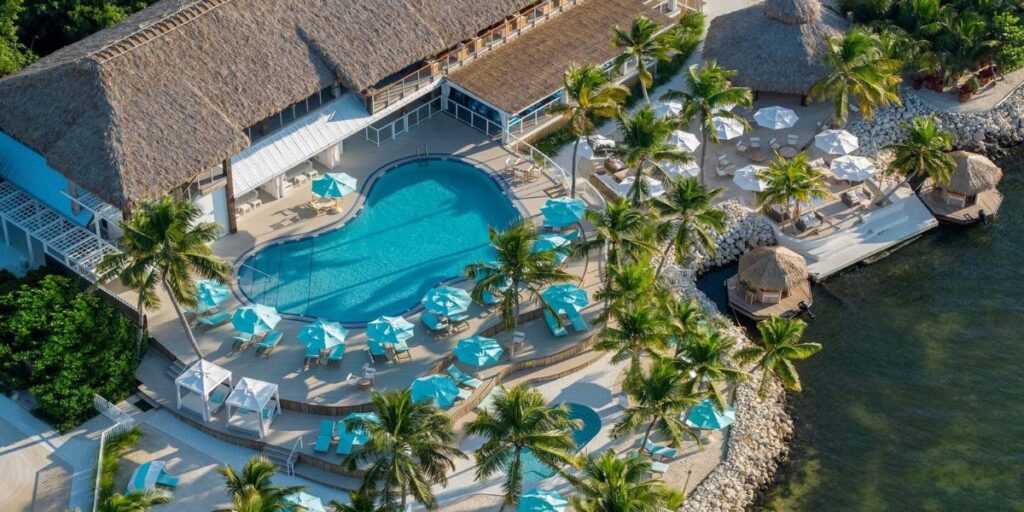 Aerial view of a luxury hotel poolside with cabanas, palm trees, and loungers, adjacent to a calm sea.