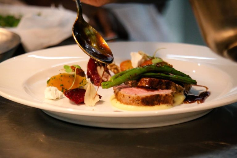 A chef drizzles sauce over a gourmet plated dish featuring sliced meat and vegetables.