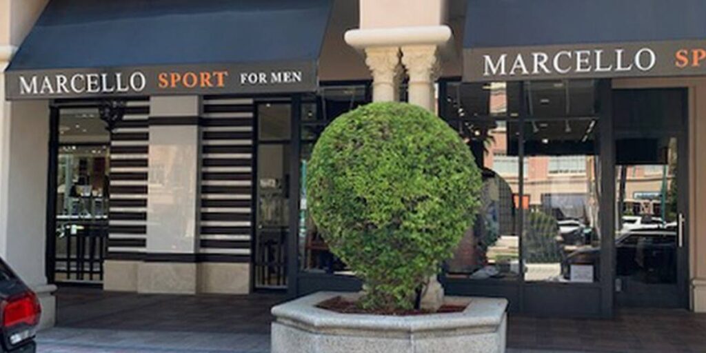A trimmed green shrub sculpted into a spherical shape sits in a stone planter in front of the Marcello Sport for Men clothing store.