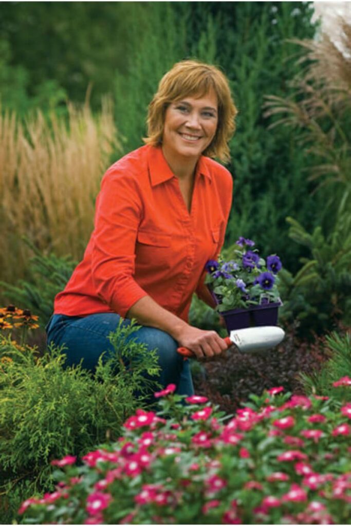 Melinda Myers smiling in a garden while holding a pot of purple flowers, with lush greenery and pink blossoms in the background.