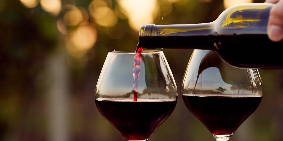 Red wine being poured into two glasses against a soft-focus background with warm sunlight.