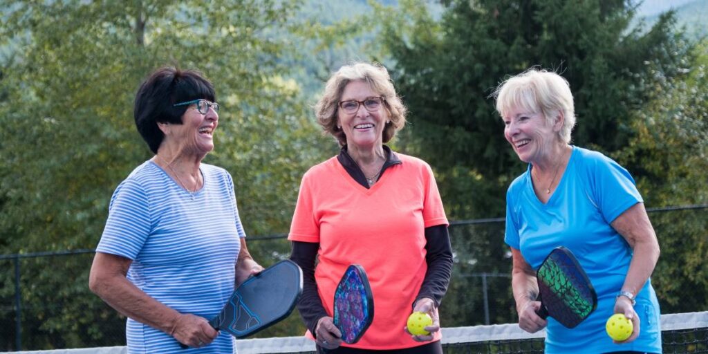 Three elderly women sharing a laugh on a pickleball court, holding paddles and a ball.