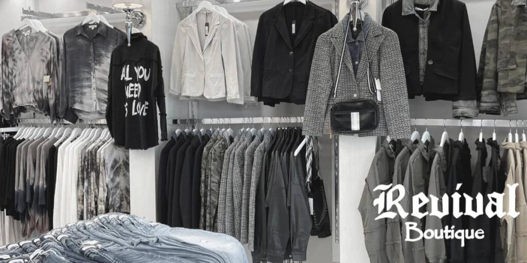 A trendy display at Revival Boutique featuring an array of monochromatic and tie-dye clothing, stylish jackets, and casual jeans, highlighted by a central black shirt with the text "ALL YOU NEED IS LOVE" in a white font.