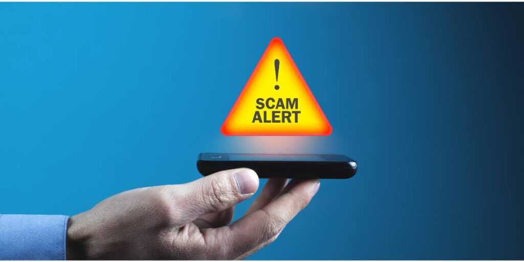 Hand holding a smartphone with a scam alert warning icon floating above it against a blue background.