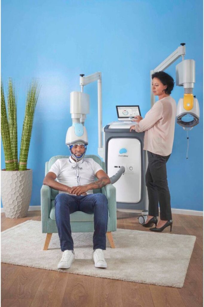 A patient undergoing transcranial magnetic stimulation (TMS) treatment while seated comfortably in a chair, as a healthcare provider operates the TMS machine.