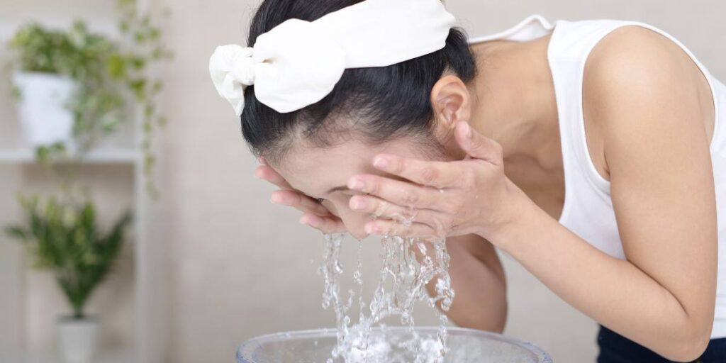 A woman with a headband washing her face with water over a basin, with indoor plants in the background.