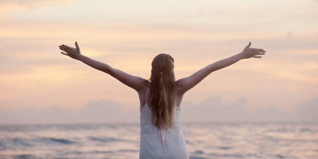 A woman with arms wide open facing a sunset over the ocean, symbolizing freedom and happiness.