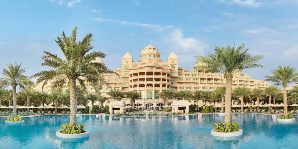  A palatial resort with a massive central swimming pool surrounded by palm trees.