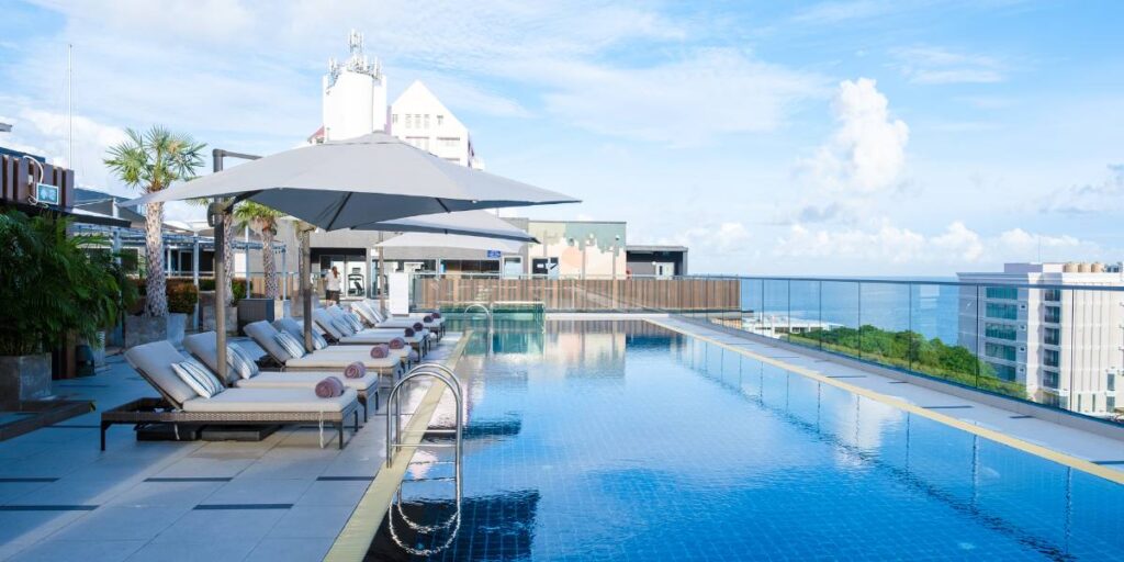 A rooftop infinity pool with lounge chairs and umbrellas, overlooking a coastal cityscape.