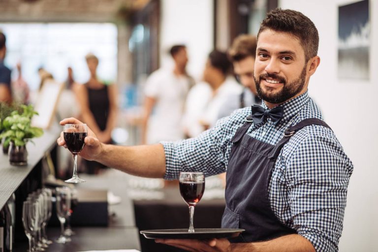 A cheerful waiter presenting a glass of red wine on a tray in a bustling restaurant